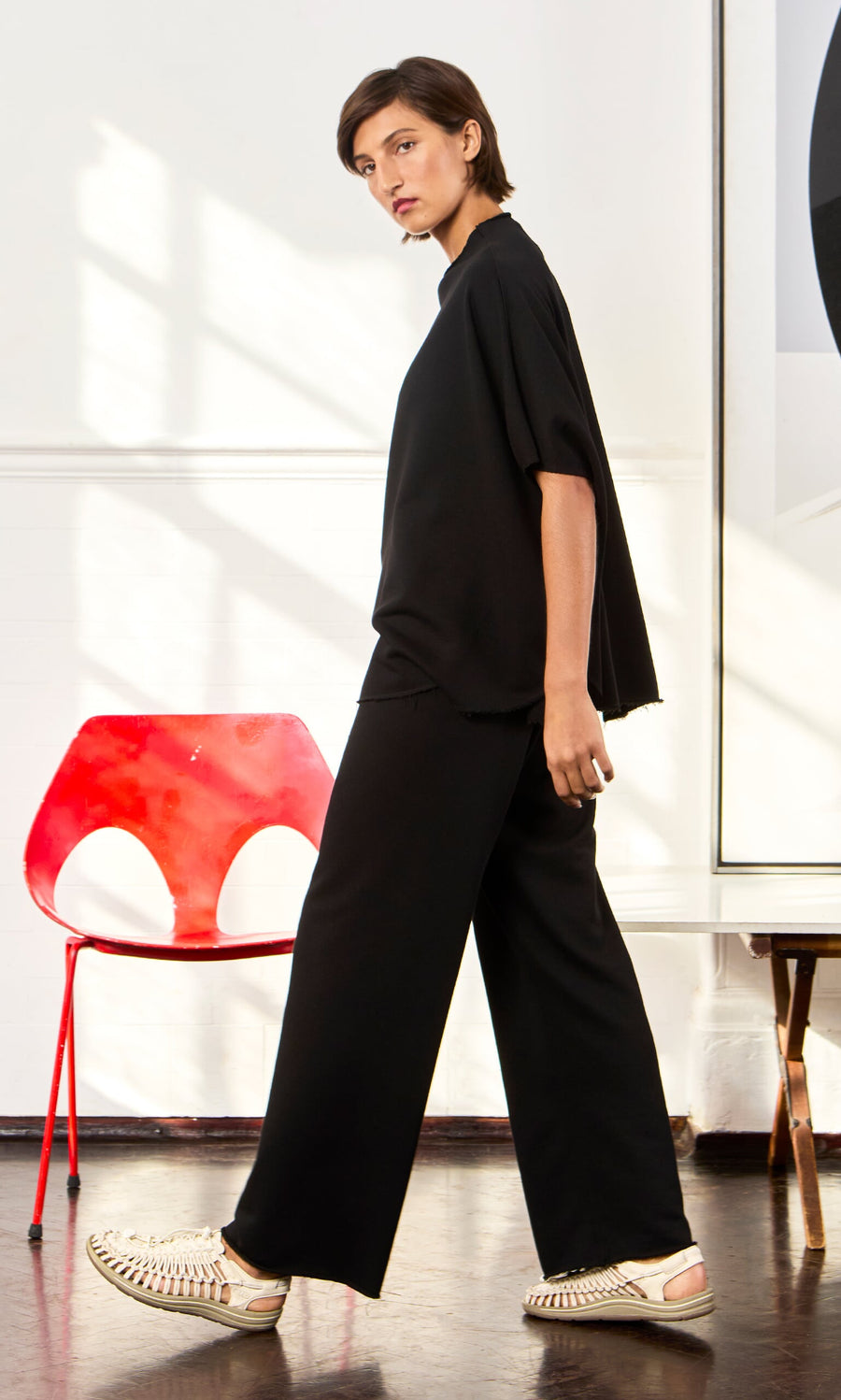 Plümo- Person in a Gabriella Top and woven shoes walks past a modern red chair in a bright, sunlit room.