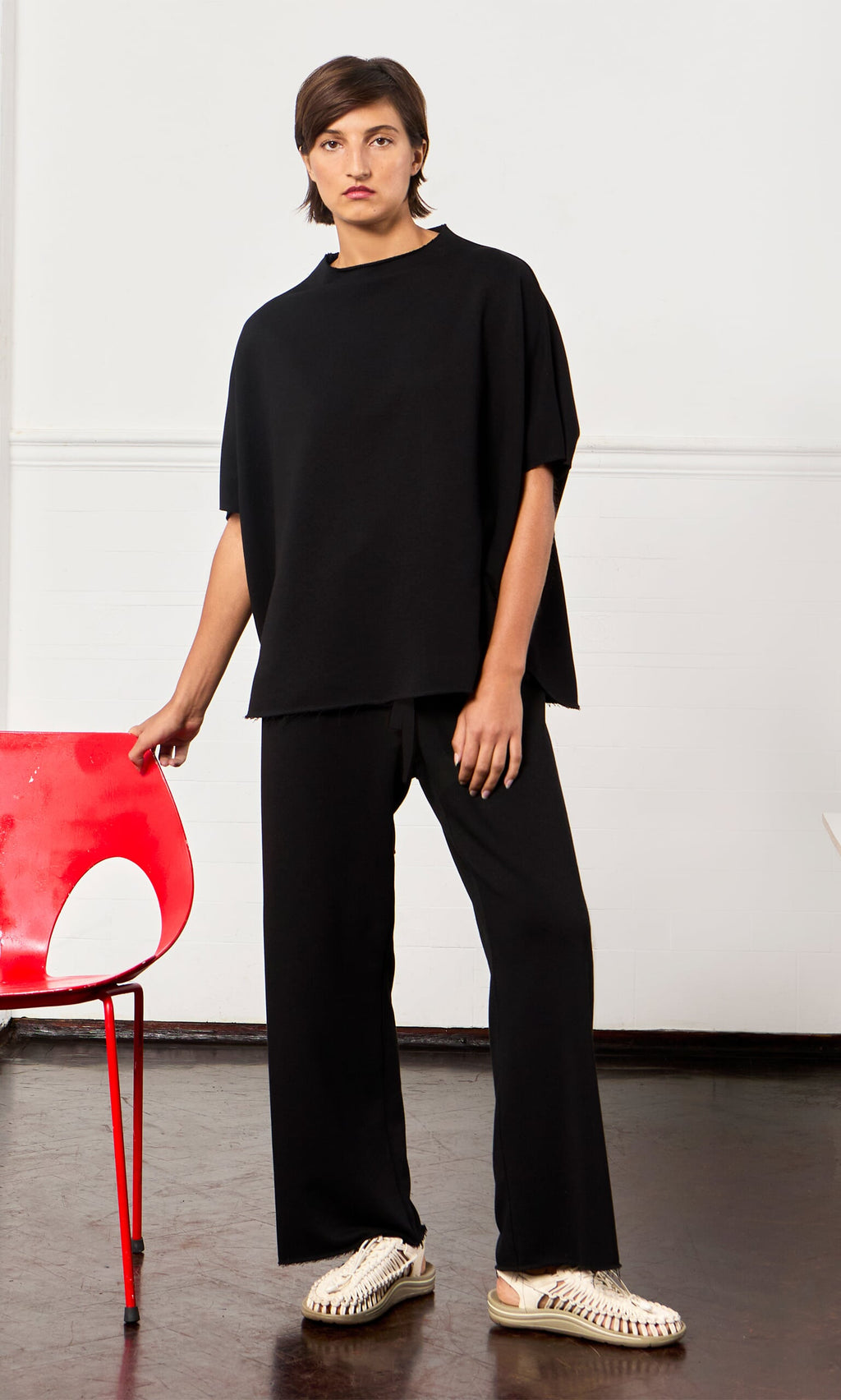 Plümo- A person in the Gabriella Top and pants stands by a red chair, wearing beige sandals in a minimalist room.