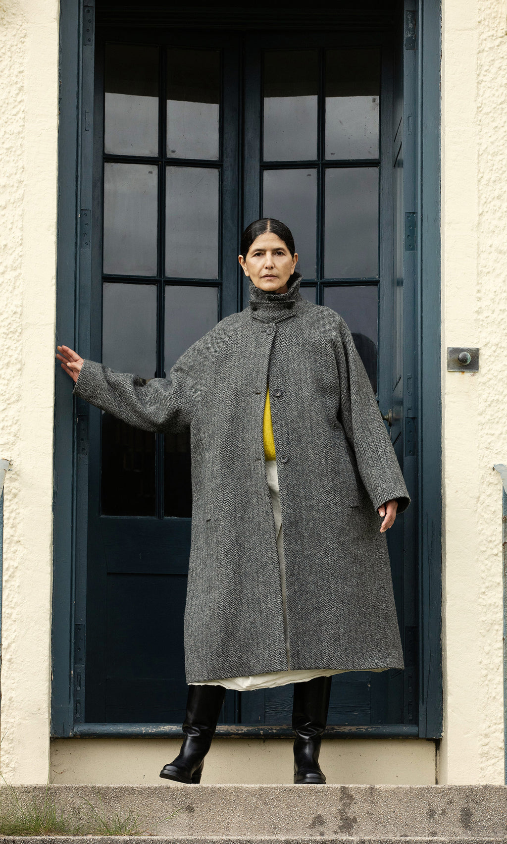 Plümo  A person in a Balmacaan Tweed Coat stands on steps before a blue door, one arm outstretched. Made in England.