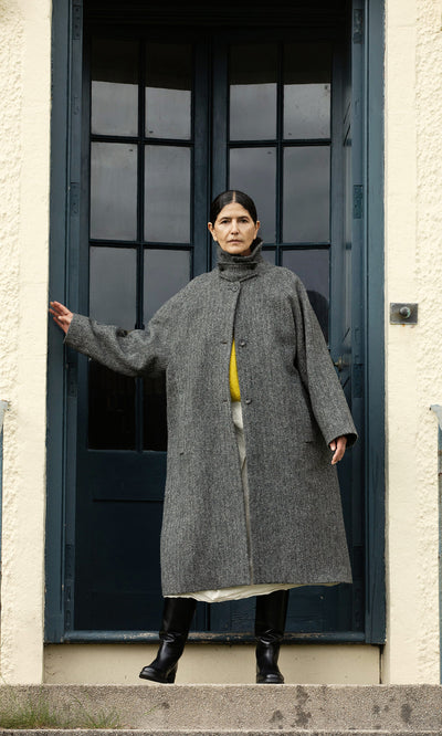 Plümo  A person in a Balmacaan Tweed Coat stands on steps before a blue door, one arm outstretched. Made in England.