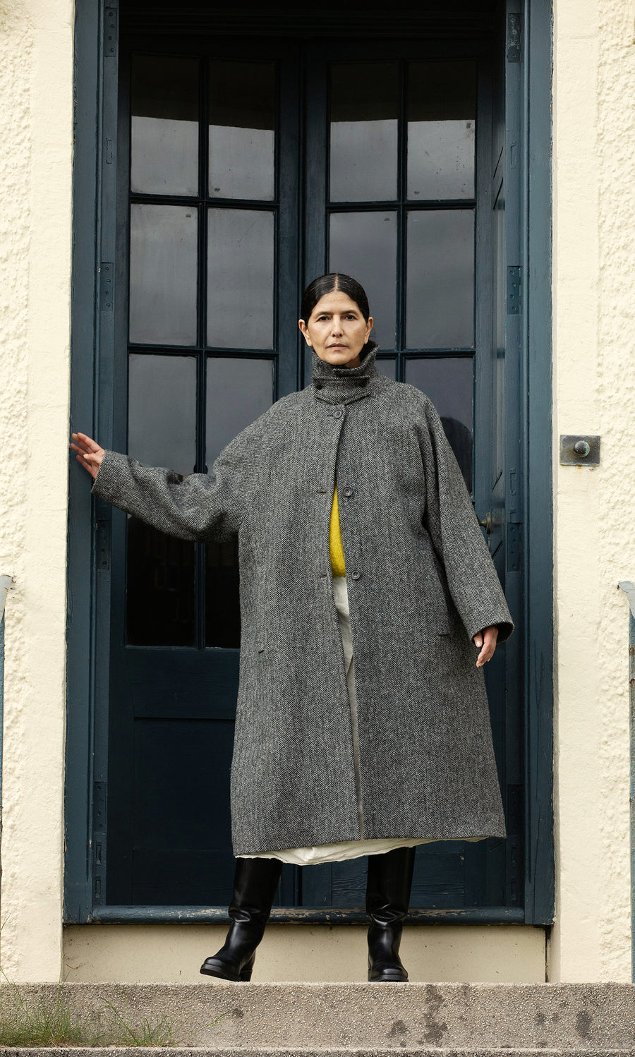 Plümo  A person in a Balmacaan Tweed Coat stands on steps before a blue door, one arm outstretched. Made in England.