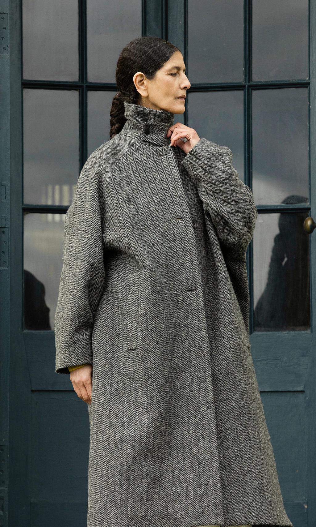 Plümo  A woman in a Balmacaan Tweed Coat stands by blue doors, gazing aside with one hand touching her collar.