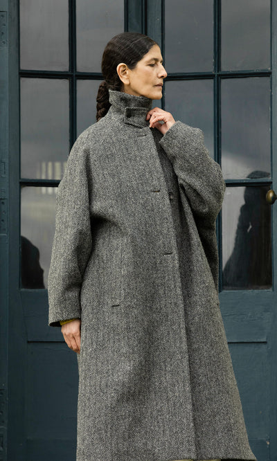 Plümo  A woman in a Balmacaan Tweed Coat stands by blue doors, gazing aside with one hand touching her collar.