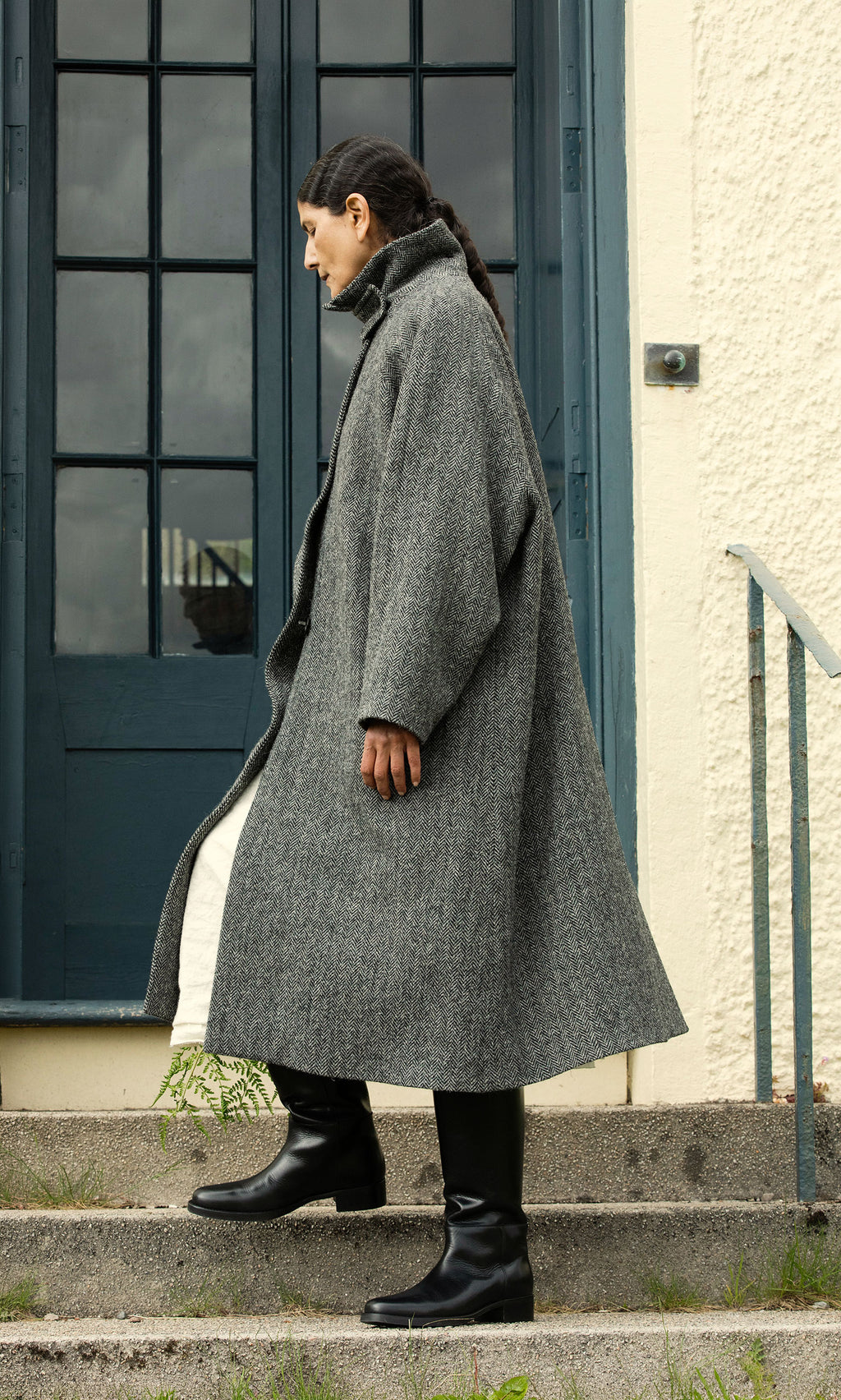 Plümo  Someone in a Balmacaan Tweed Coat and black boots stands sideways on concrete stairs by a blue door.
