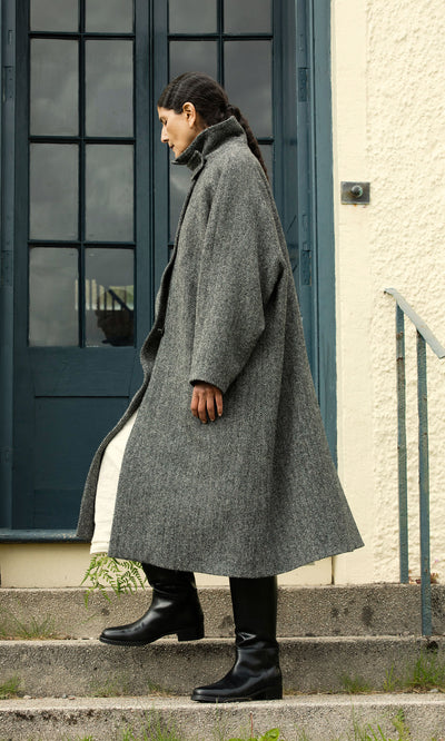 Plümo  Someone in a Balmacaan Tweed Coat and black boots stands sideways on concrete stairs by a blue door.