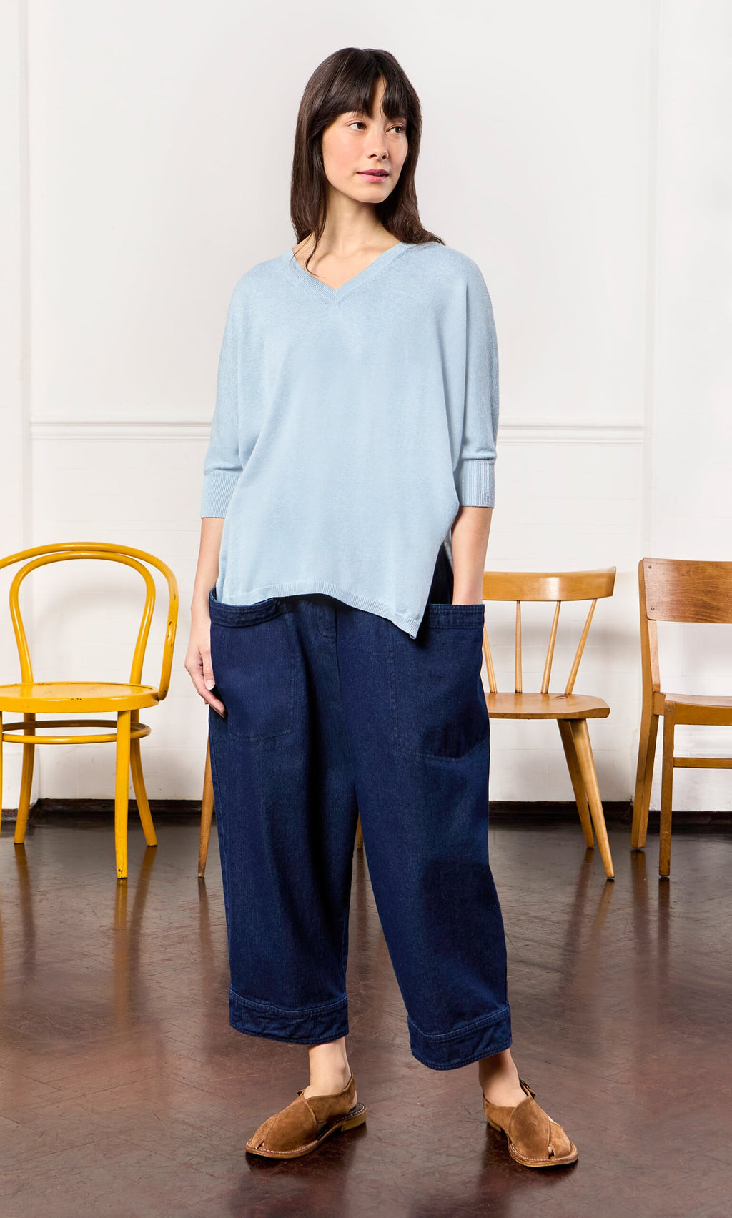 Plümo- Woman in a Batwing Jumper, wide dark blue pants, and brown slippers stands near three wooden chairs.