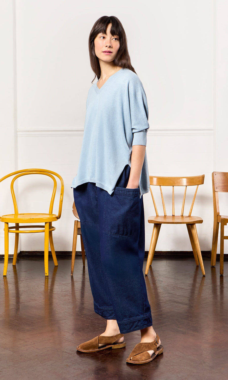 Plümo- Woman in a Batwing Jumper relaxed fit light blue top and dark blue pants stands indoors; wooden chairs behind her.