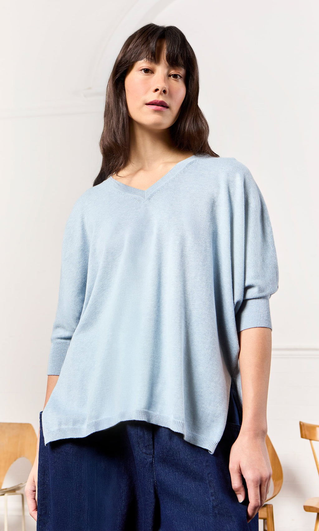 Plümo- Woman in a relaxed fit light blue Batwing Jumper and dark pants, standing indoors with chairs in the background.