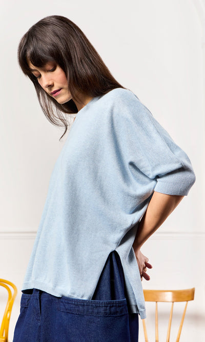 Plümo- Woman in a relaxed fit Batwing Jumper and dark pants stands sideways, looking down by a wooden chair.