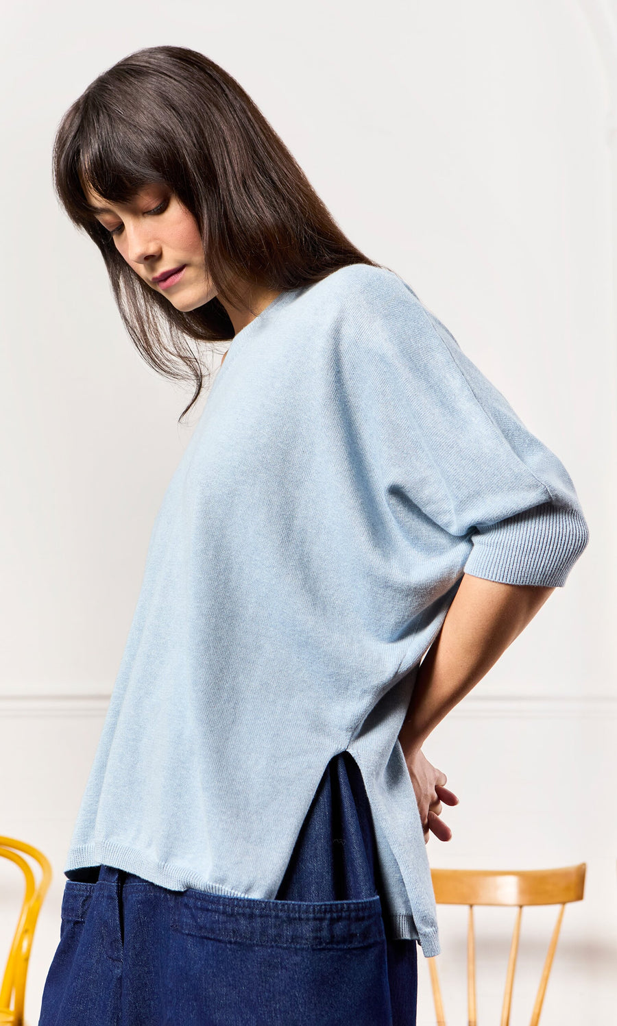 Plümo- Woman in a relaxed fit Batwing Jumper and dark pants stands sideways, looking down by a wooden chair.