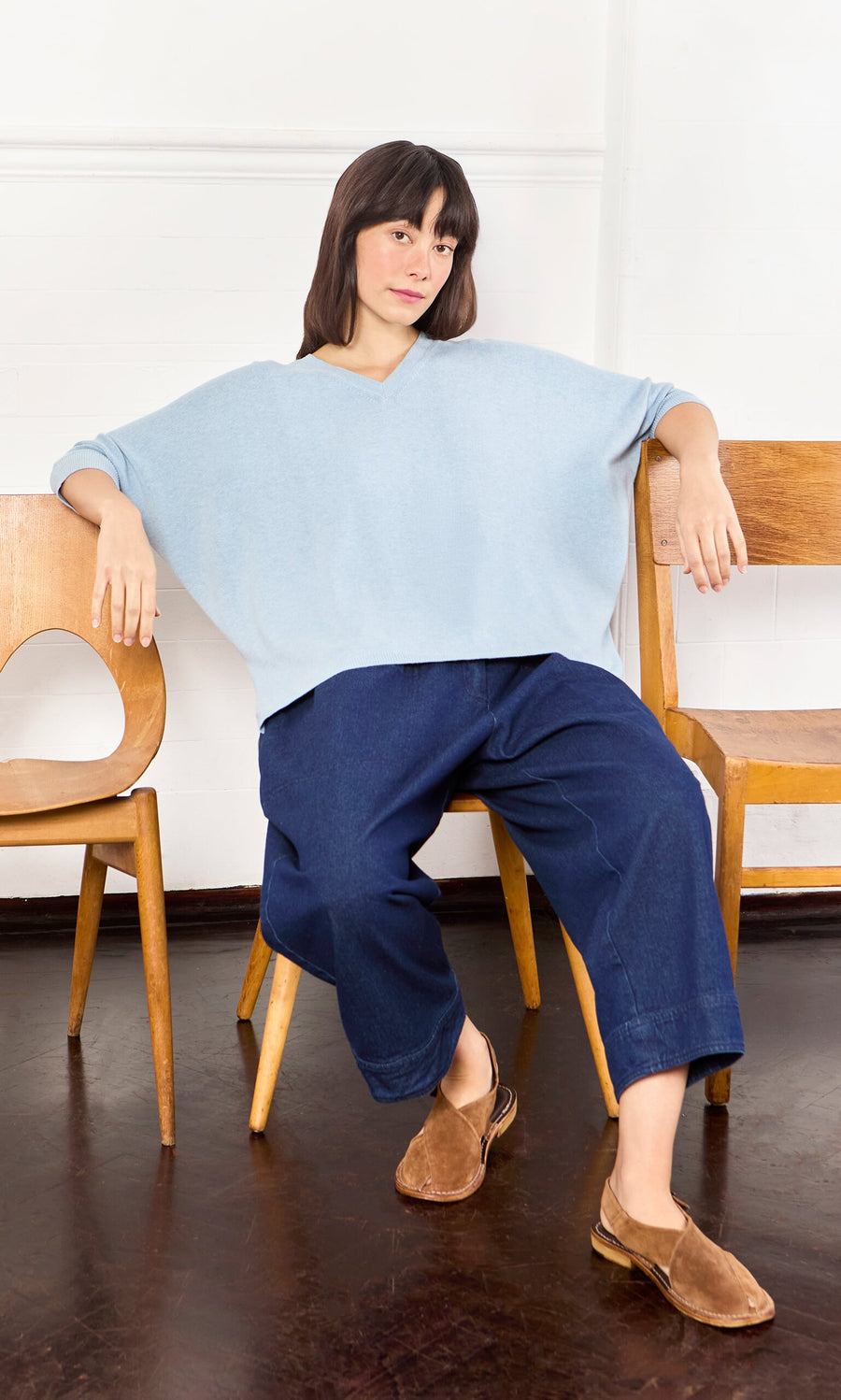 Plümo- Woman in a Batwing Jumper and dark blue pants sits on a wooden chair between two others indoors.
