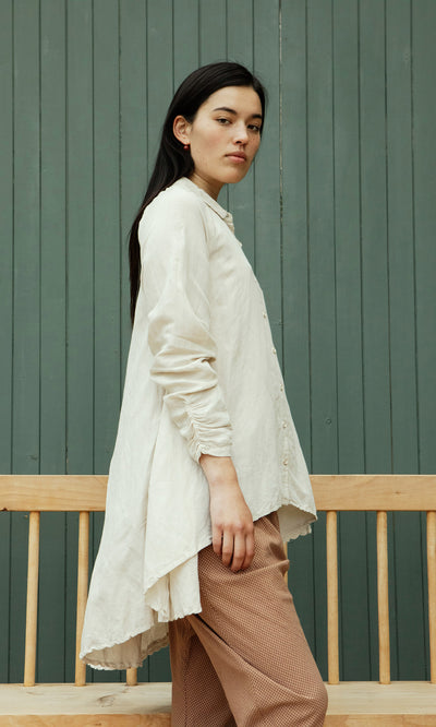 Plümo  A woman in the Beeson Shirt - Oat and brown pants stands sideways by a wooden bench, before green paneled doors.