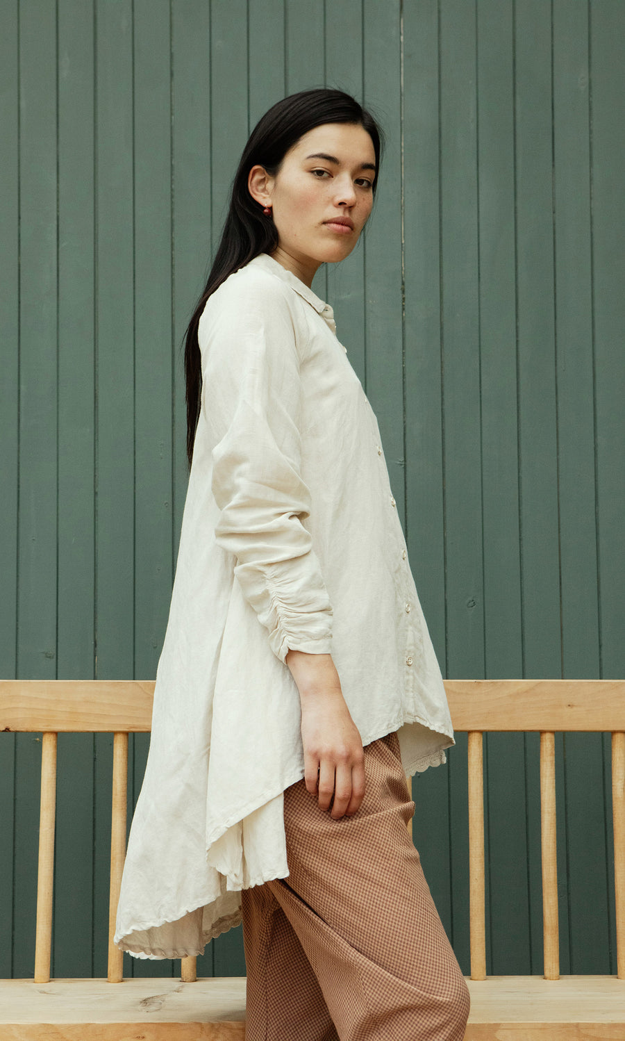 Plümo  A woman in the Beeson Shirt - Oat and brown pants stands sideways by a wooden bench, before green paneled doors.