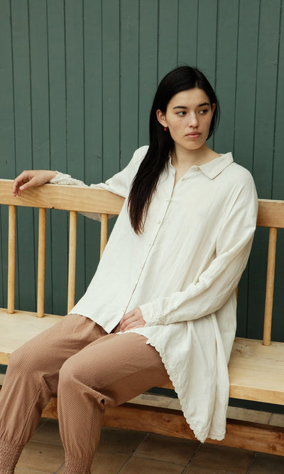 Plümo  Woman in Beeson Shirt - Oat and brown pants sits on a bench against a green paneled wall.