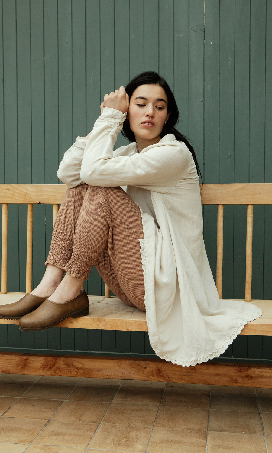Plümo  Woman in the Beeson Shirt - Oat and brown pants sits sideways on a wooden bench by a green paneled wall.
