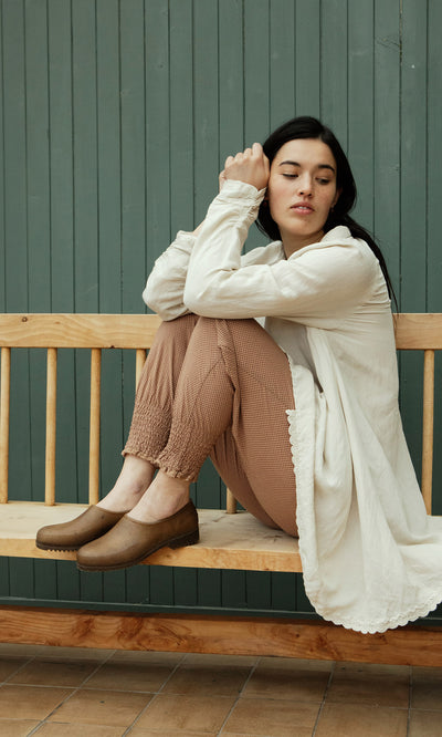 Plümo  Woman in a white blouse and brown pants wears Hemp Clogs, sitting on a bench by a green paneled wall.