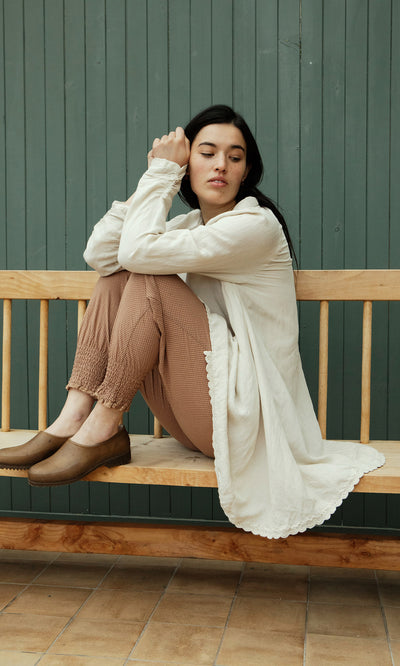 Plümo  Woman in cream cardigan and Vichy Pantaloons sits sideways on a wooden bench by a green paneled wall.