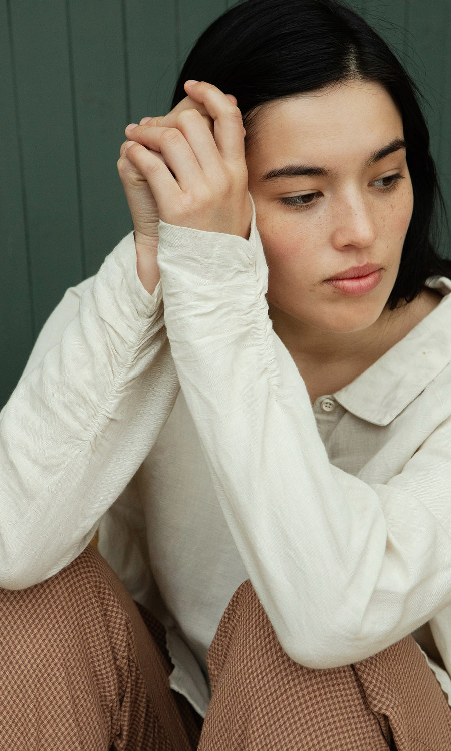 Plümo  A person with dark hair sits, hands clasped, wearing the Beeson Shirt - Oat and brown pants, looking thoughtfully aside.