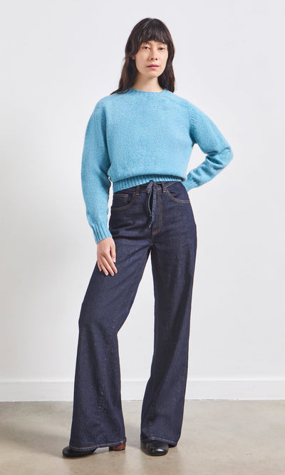 Plümo- Person standing in a light blue sweater and Belted Wide Jeans against a plain white background.