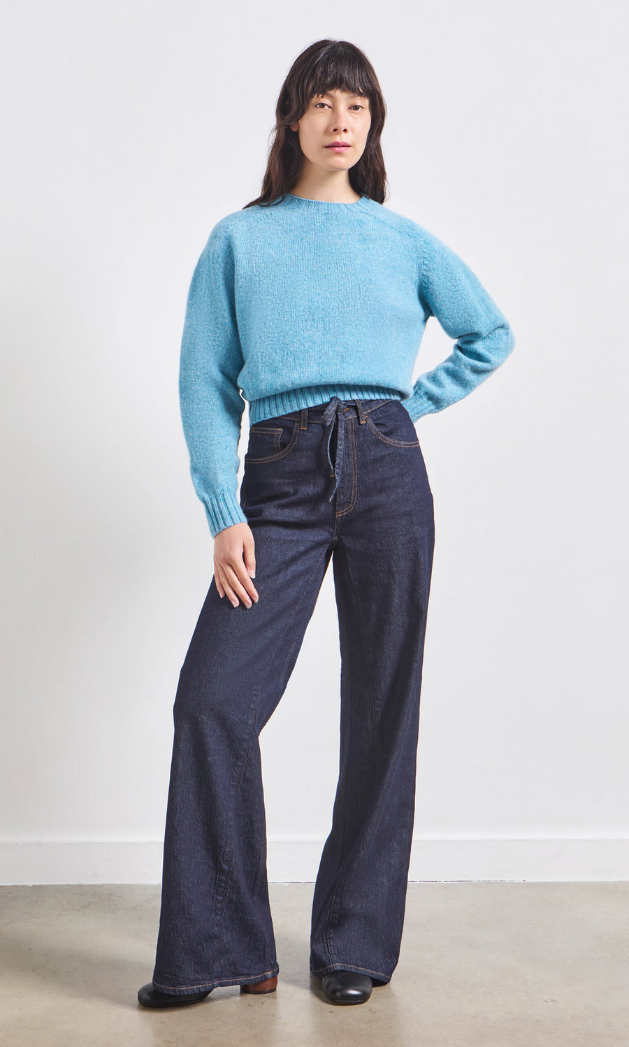 Plümo- Person standing in a light blue sweater and Belted Wide Jeans against a plain white background.