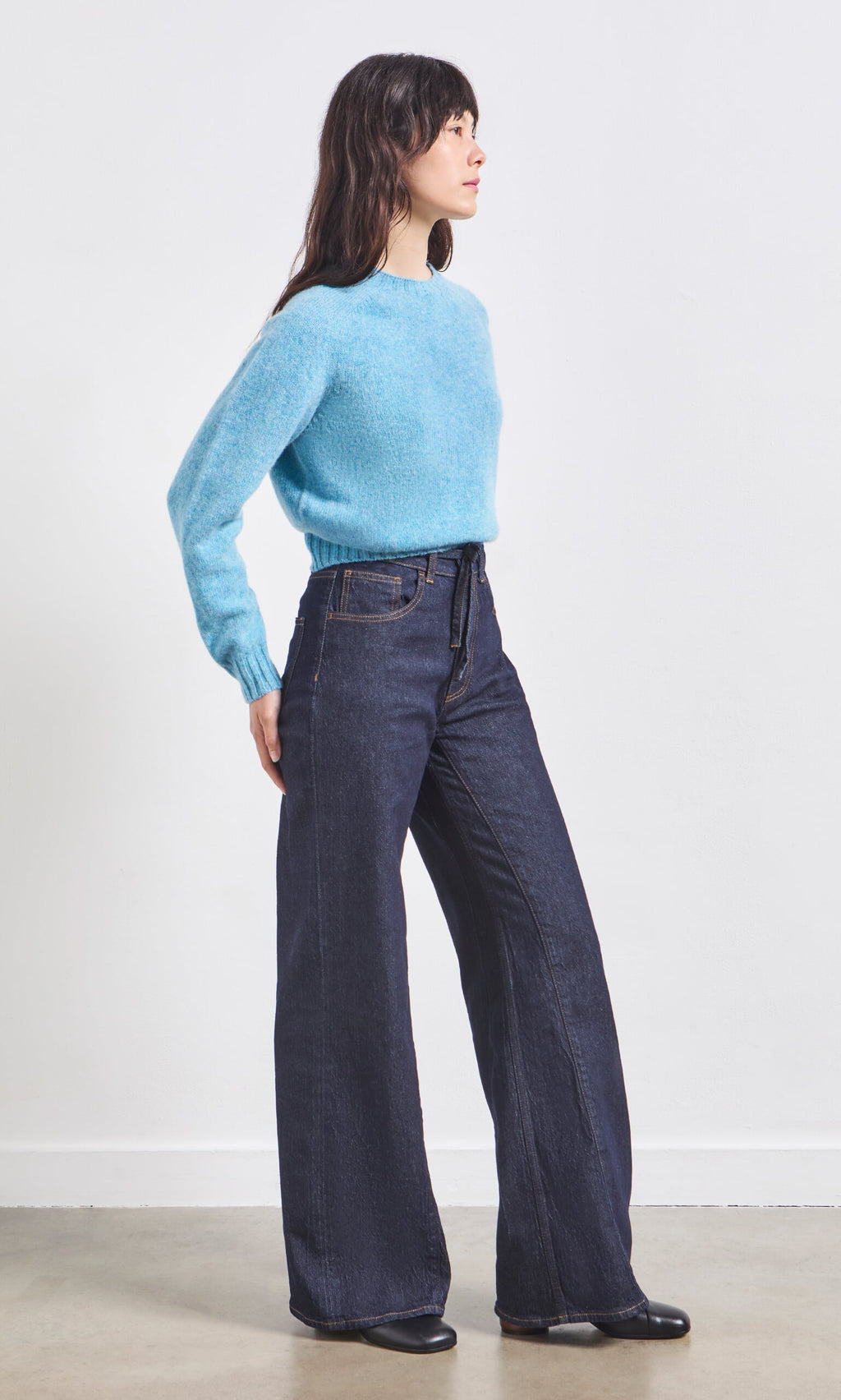 Plümo- Woman in a light blue sweater and Belted Wide Jeans stands sideways against a plain white wall.