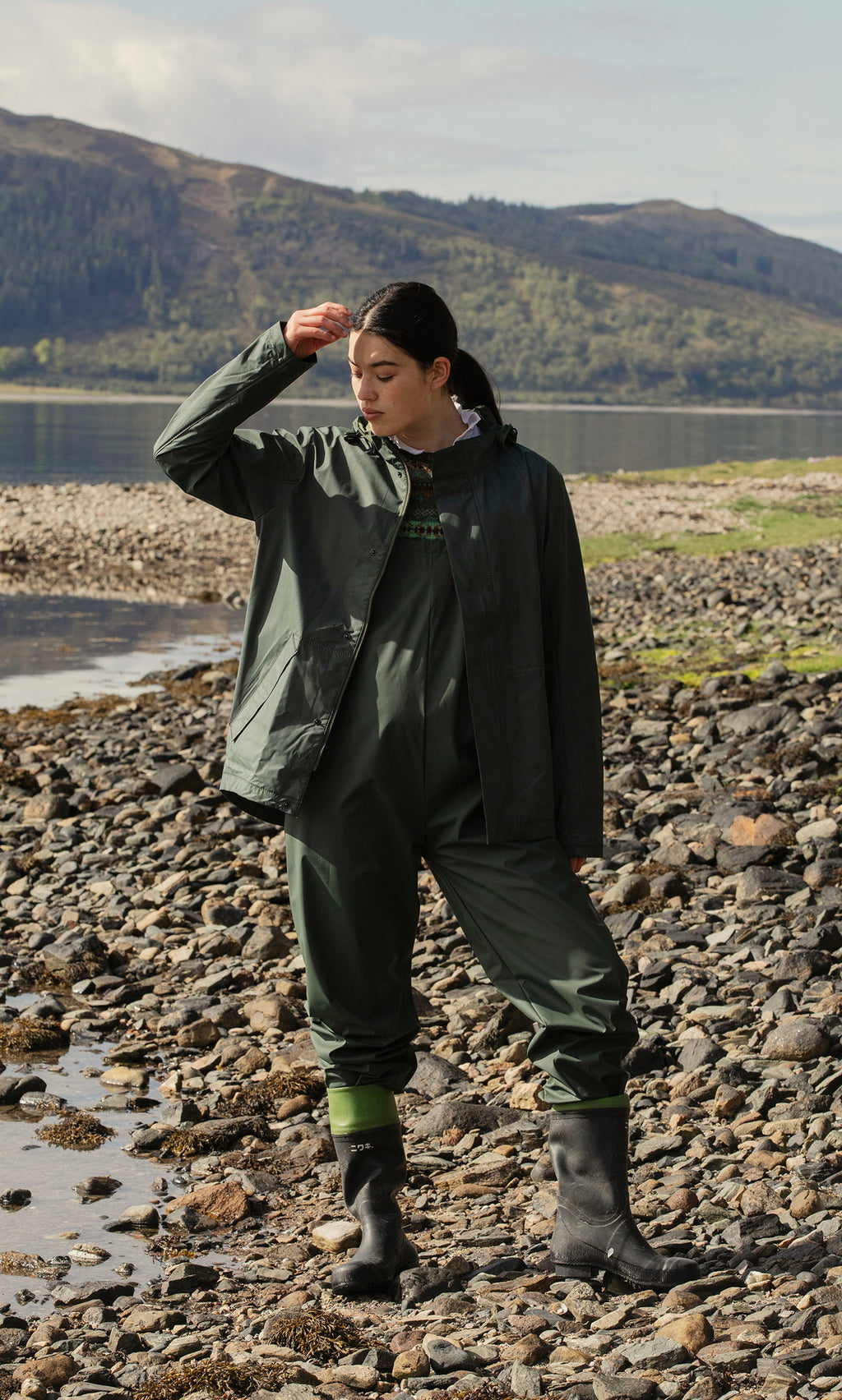 Plümo  A person in green Land Dungarees and boots stands on lakeshore rocks, touching their cap with hills behind.