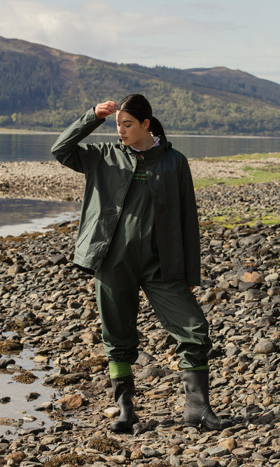 Plümo  A person in green Land Dungarees and boots stands on lakeshore rocks, touching their cap with hills behind.