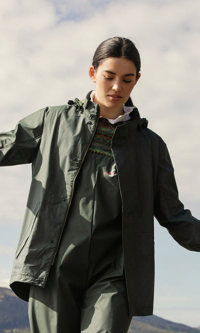 Plümo  Person outdoors in a green Berghart Raincoat, looking down, with cloudy sky and hills in the background.
