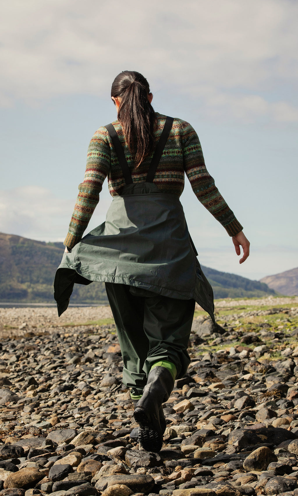Plümo  Person in Land Dungarees and sweater walks on rocky shore, facing away, with hills and sky in the background.