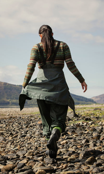 Plümo  Woman in green waders and Berghart Raincoat walks on rocky ground near hills, photographed from behind.