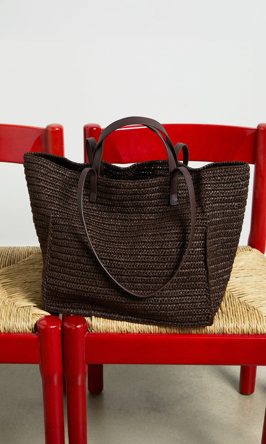 Plümo- The Tova Bag, a dark brown straw tote with leather handles, rests on two red chairs with woven straw seats.