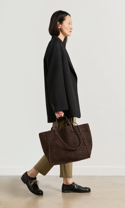 Plümo- Woman in a black blazer and khaki pants carrying the boxy Tova Bag, a large straw tote, walking sideways.