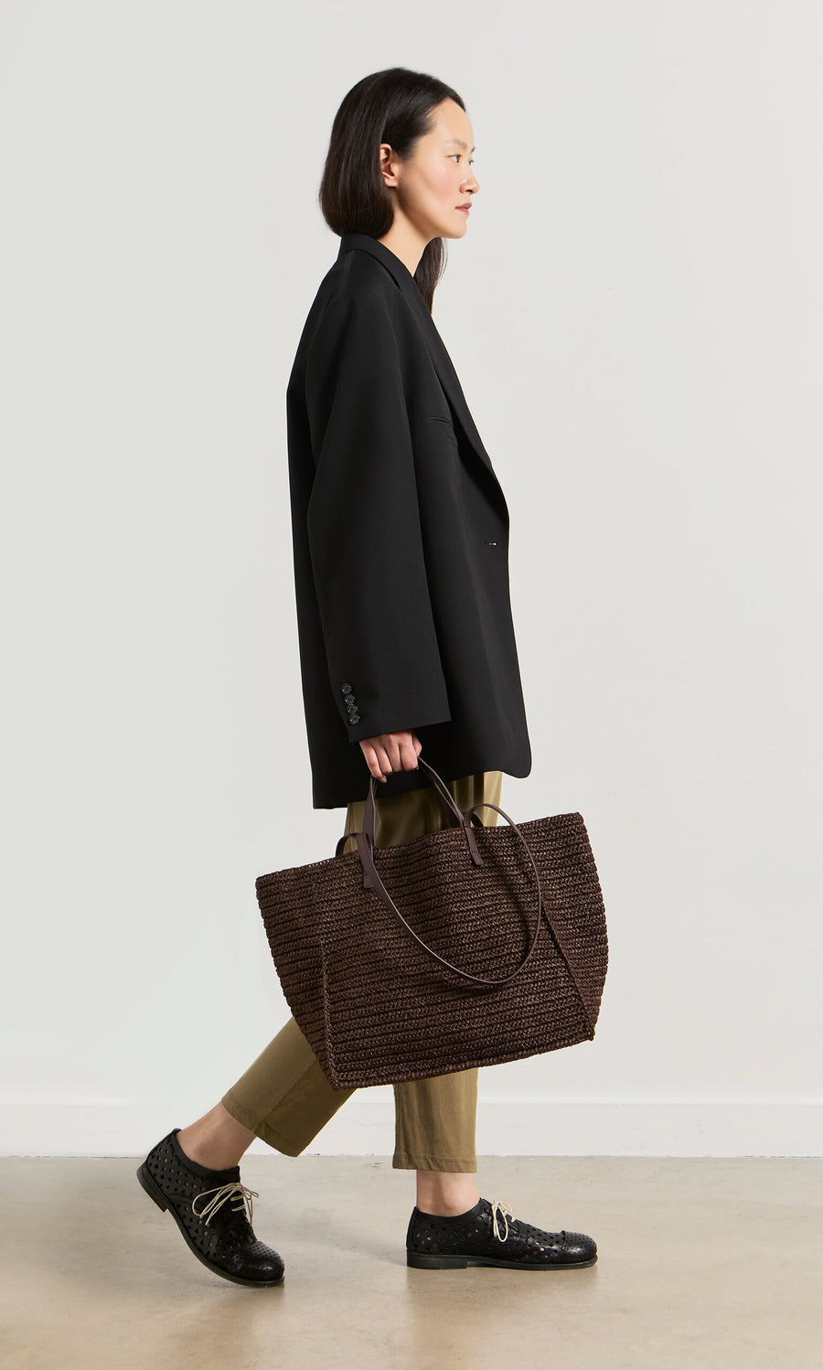 Plümo- Woman in a black blazer and khaki pants carrying the boxy Tova Bag, a large straw tote, walking sideways.