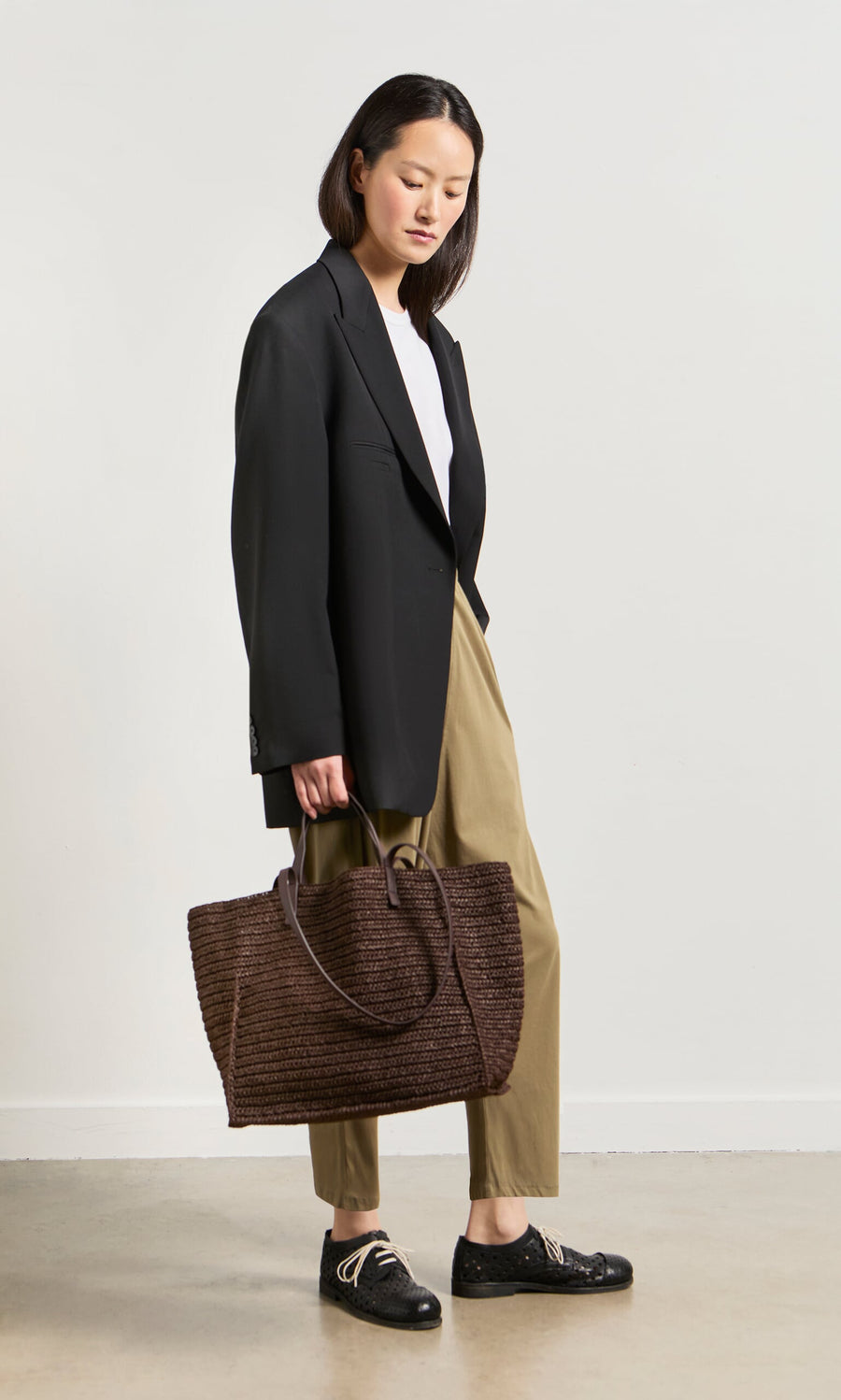 Plümo- Woman in a black blazer, white top, khaki pants, black shoes, holding the boxy-structured Tova Bag straw tote.