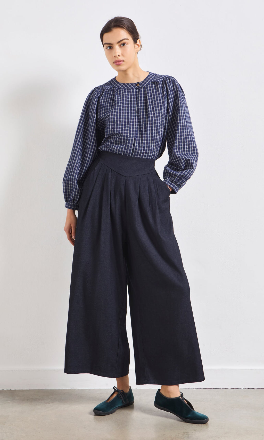 Plümo- Woman wearing the Little Prairie Blouse and wide-leg navy pants stands against a white wall, looking at the camera.