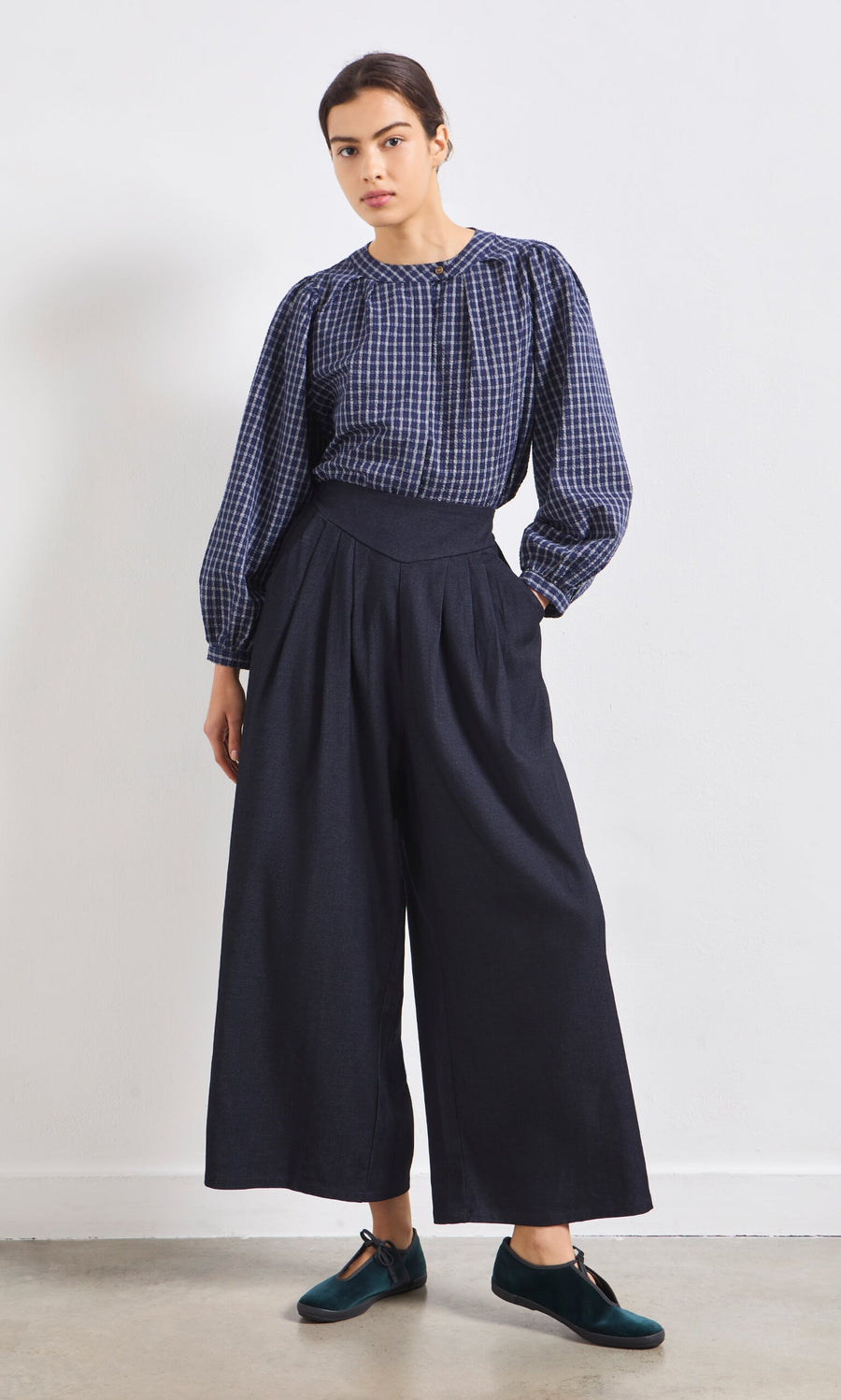 Plümo- Woman wearing the Little Prairie Blouse and wide-leg navy pants stands against a white wall, looking at the camera.
