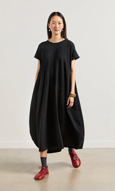 Plümo- Woman in the Blackout Dress, a relaxed-fit stretch jersey, stands indoors against a plain light-colored wall.
