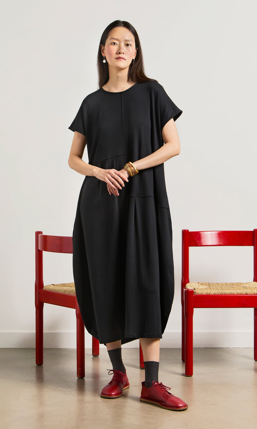 Plümo- Woman in a relaxed fit Blackout Dress and red shoes stands between two red chairs against a white wall.