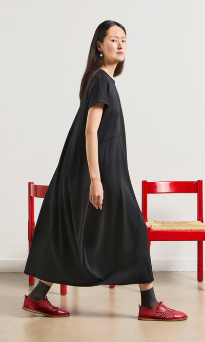 Plümo- Woman in a relaxed fit Blackout Dress and red shoes walks past two red chairs against a white wall.