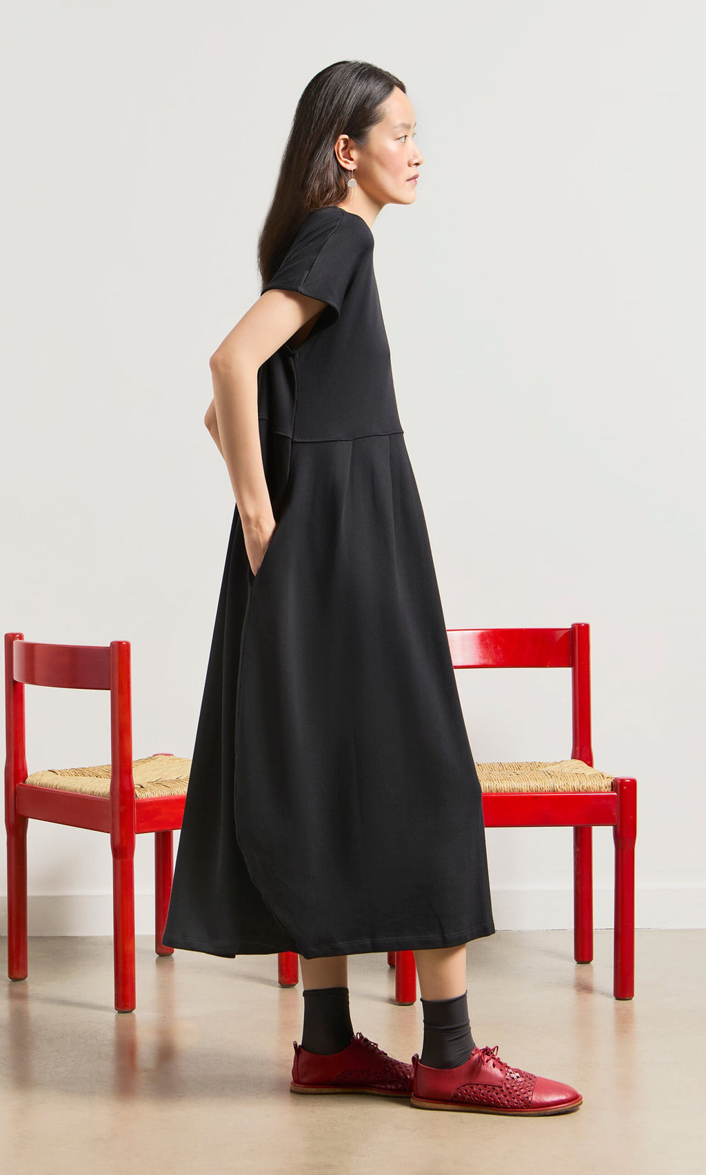 Plümo- Woman in the Blackout Dress and red shoes stands sideways by two red chairs on a neutral background.