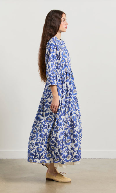 Plümo- Woman in the Blue Pansy Dress and beige shoes stands sideways against a plain white wall.