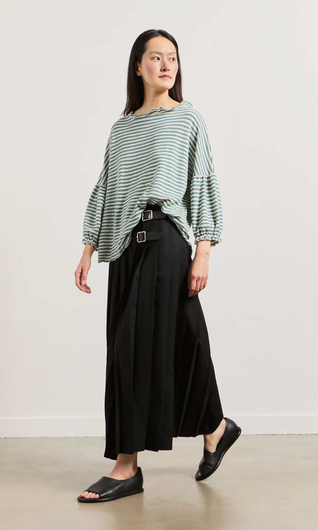 Plümo- A woman in a Blue Stripe Ruffle Blouse, black pleated skirt with belts, and black slides stands against a white background.