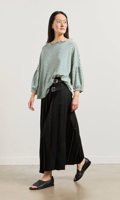 Plümo- A woman in a Blue Stripe Ruffle Blouse, black pleated skirt with belts, and black slides stands against a white background.