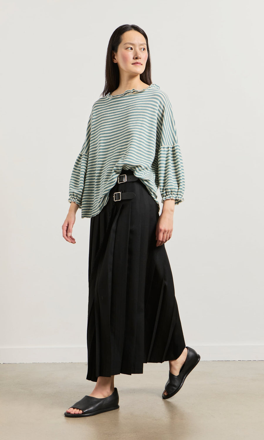 Plümo- A woman in a Blue Stripe Ruffle Blouse, black pleated skirt with belts, and black slides stands against a white background.