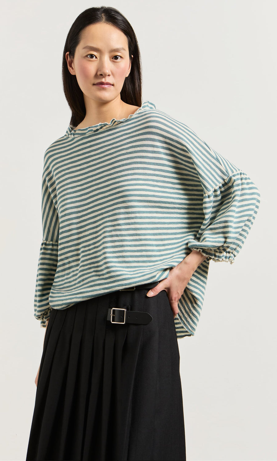 Plümo- Woman wearing a Blue Stripe Ruffle Blouse and black pleated skirt with a belt, standing against a plain background.