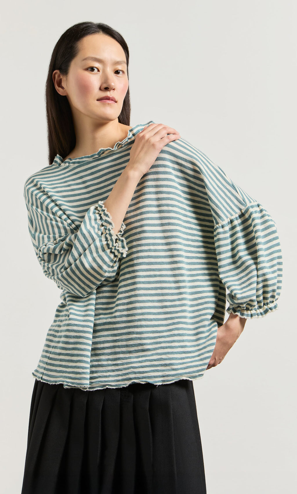 Plümo- Woman wears a Blue Stripe Ruffle Blouse with puffed sleeves, paired with a black skirt against a plain background.
