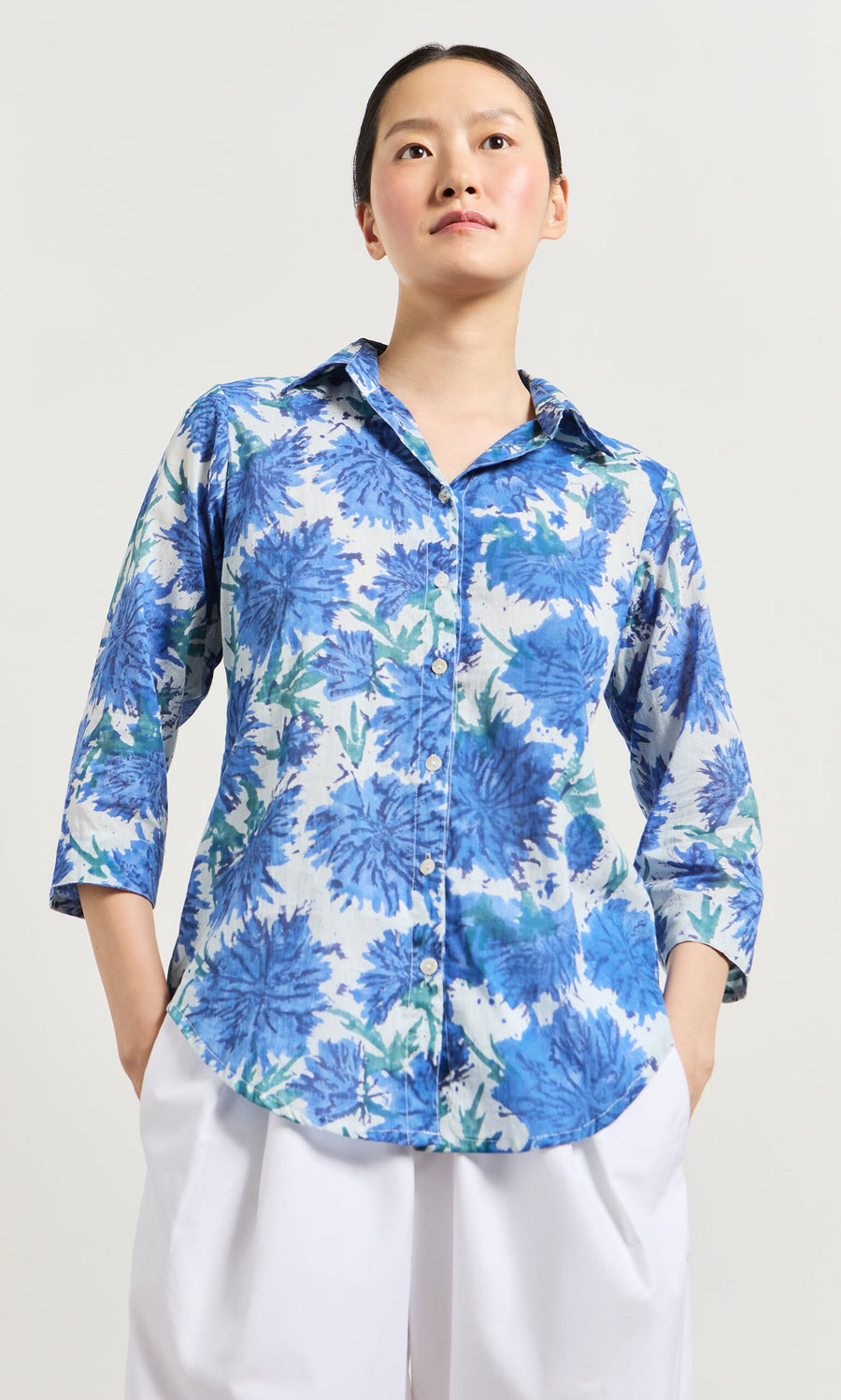 Plümo- A woman wears the Blue Thistle Shirt with white pants, standing with hands in pockets against a plain background.
