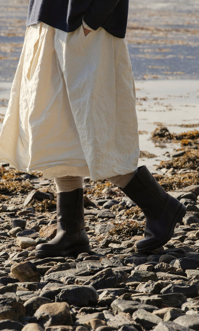Plümo  Person in a cream skirt and Rigger Boots walks on rocky ground by water, with seaweed in the background.