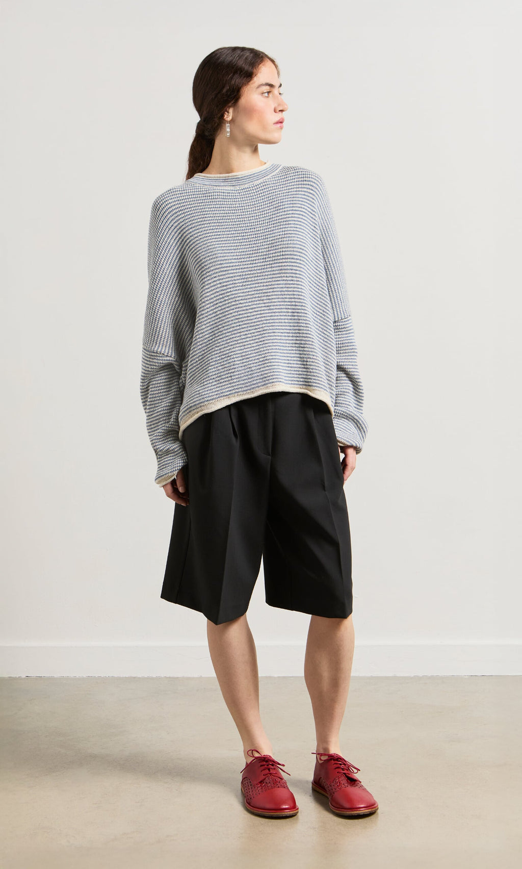 Plümo- Woman in a Boathouse Jumper of natural cotton yarns, black shorts, and red shoes stands facing left.