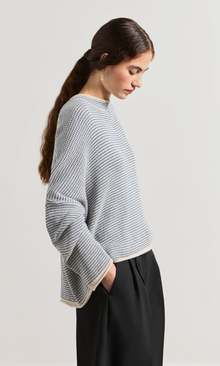 Plümo- Woman with long brown hair in a ponytail wears the Boathouse Jumper and black pants, looking downward.